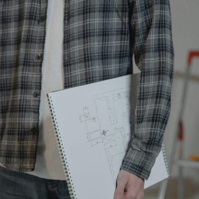 Man holding a sketchbook with architectural blueprints indoors, wearing a plaid shirt.